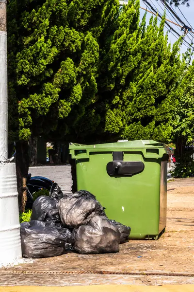 Yeşil çöp konteynırları ve siyah çöp torbaları yapraklı bir sokak kaldırımında. Atık yönetimi, kentsel hijyen ve çevresel farkındalık.