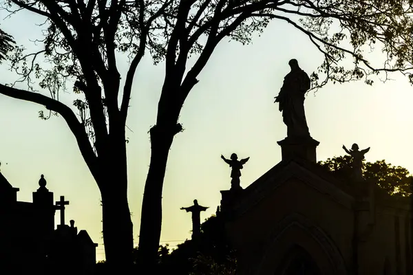 Mezarlık siluetleri alacakaranlıkta, renkli gökyüzüne karşı haçlar ve heykellerle. İnanç, hafıza ve yansıma temalarını çağrıştırır