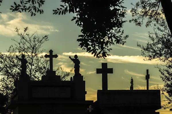 Mezarlık siluetleri alacakaranlıkta, renkli gökyüzüne karşı haçlar ve heykellerle. İnanç, hafıza ve yansıma temalarını çağrıştırır