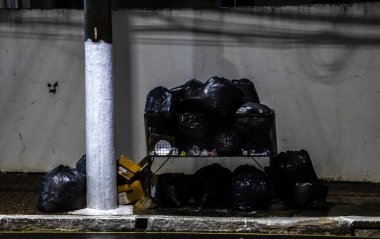 Sao Paulo, Brezilya, 23 Haziran 2025. Metal bir çöp konteynırından taşan siyah çöp torbaları beton direğinin yanındaki beyaz bir duvara çarpıyor. Gece sahnesinde kentsel atıklar ve çevre sorunları vurgulanıyor.