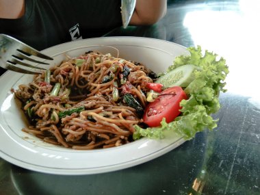 fried noodles with lettuce, tomato, cucumber on a white plate with a wooden background