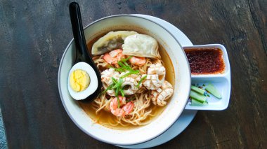 Bir kase acılı erişte çorbası ve ahşap bir masada salatalık turşusu..