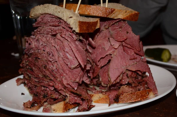 A tall corned beef and pastrami deli sandwich on rye bread on a white plate, A pickle can be seen in the background