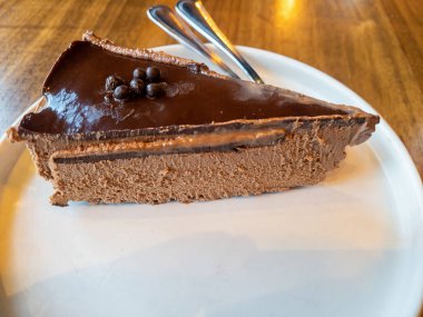 Beyaz tabakta çikolatalı cheesecake. Ahşap bir masada. Arka planda gümüş takımlar var..
