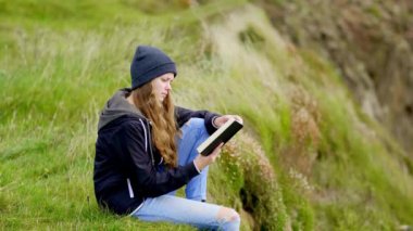 Genç bir kadın İrlanda 'da uçurumun kenarında otururken kitap okuyor. Ağır çekim klibi. 