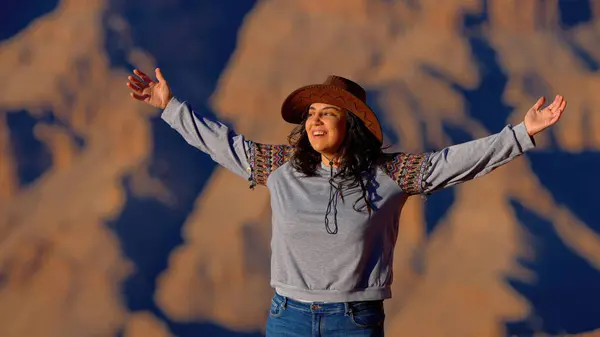 A free-spirited cowgirl stands proudly at the edge of the grand canyon, her jeans blending with the rugged desert landscape as the fiery sunset illuminates her outstretched arms and her soul connects