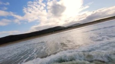 Sahil üzerinde alçak ve yüksek hızlı FPV uçuşu ve okyanus suyu okyanus dalgalarının sakin ve sakin görüntüsü parlak mavi gökyüzünün altında yavaşça kıyıya çarpıyor.