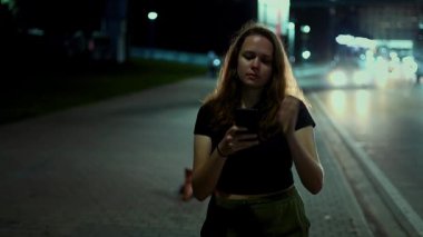 Genç bir kadın telefonuna derinden odaklanmış. Etrafı canlı şehir ışıkları ve gece hareket eden araçlarla çevrili.