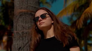 Şık bir kadın, şık güneş gözlükleriyle güneşli tropikal atmosferin tadını çıkararak uzun bir ağaca yaslanıyor.