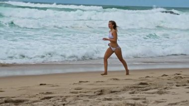Dalgalar arka planda güçlü bir şekilde düşerken kumsalda canlı canlı koşan neşeli genç bir kadın.
