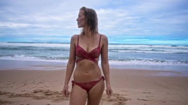 Çekici bir kadın sahilde duruyor, kırmızı bir bikini giyiyor, arkasında okyanus dalgaları yavaşça çarpıyor..