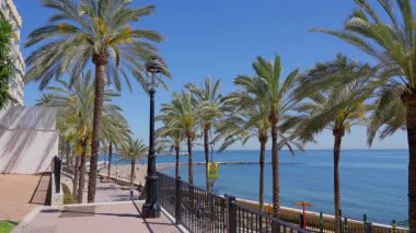 Güneşli bir yaz gününde İspanya 'nın Marbella kentindeki Akdeniz manzaralı palmiye ağaçlarıyla kaplı gezinti güvertesinde yürüyen turistler - MARBELLA, İspanya - 30 Mart 2025