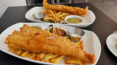 Lezzetli balık ve patates kızartması beyaz tabakta soslu ve limonlu siyah bir masada servis ediliyor.