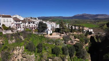 İspanya 'nın Ronda kentinin panoramik manzarasında geleneksel beyaz evler, yemyeşil bitki örtüsü ve uzaktaki Serrania de Ronda dağları yer alıyor.