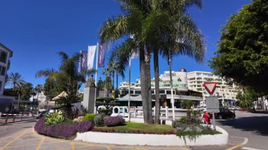 Puerto Banus, Marbella 'da döner kavşak, palmiye ağaçları, bir heykel ve açık mavi gökyüzünün altında dalgalanan bayraklar - MARBELLA, SPAIN - 30 Mart 2025