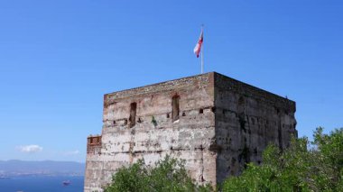 Mağribi Kalesi rüzgarda dalgalanan Cebelitarık bayrağını sergiliyor, mavi gökyüzü ve deniz arka planda