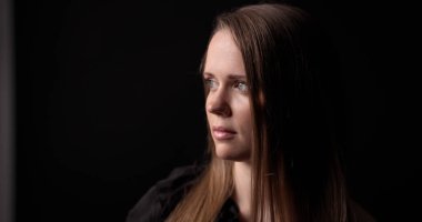 Uzun kahverengi saçlı, siyah bir arka plana bakan genç bir kadının stüdyo portresi.