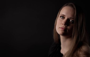 Uzun kahverengi saçlı, koyu renk elbiseli, siyah bir fonda poz veren, başı hafifçe eğilmiş ve yüzünde düşünceli bir ifade olan genç bir kadının stüdyo portresi.