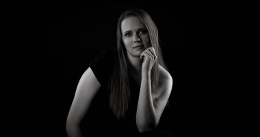 Eli çenesinde poz veren genç bir kadının stüdyo portresi, karanlık bir arka plana çarpan çarpıcı ışıkla aydınlatılmış.