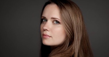 Uzun kahverengi saçlı, gri arka planda omzunun üzerinden bakan çekici genç bir kadının stüdyo portresi.