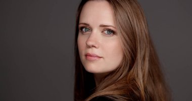 Uzun kahverengi saçlı bir iş kadınının portresi başını çeviriyor ve omuzlarının üzerinden gri bir arka plana bakıyor.