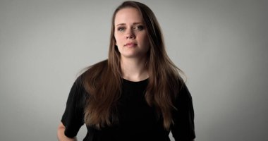 Gri arka planda uzun kahverengi saçlı, ciddi bir genç kadının stüdyo portresi.