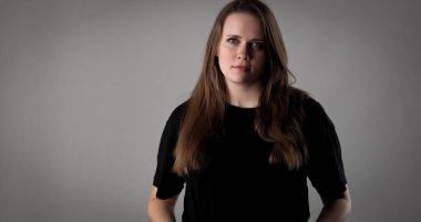 Uzun kahverengi saçlı, gri arka planda siyah tişört giyen ciddi bir genç kadının stüdyo portresi.