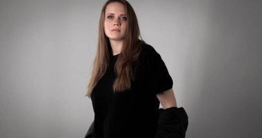 Uzun kahverengi saçlı, siyah tişört ve ceket giyen, gri arka planda poz veren genç bir kadının stüdyo portresi.