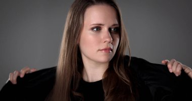 Uzun kahverengi saçlı genç bir kadının stüdyo portresi. Gri bir arkaplana bakarken siyah ceketini ayarlıyor.