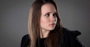 Uzun kahverengi saçlı, siyah ceketli, endişeli bir ifadeyle omzunun üzerinden bakan genç bir kadının stüdyo portresi.