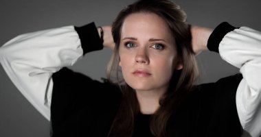 Gri arka planda elleri başının üstünde duran ciddi bir genç kadının portresi.