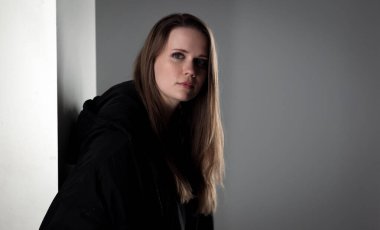 Uzun kahverengi saçlı, siyah kapüşonlu bluzlu beyaz bir duvara yaslanmış genç bir kadının stüdyo portresi.