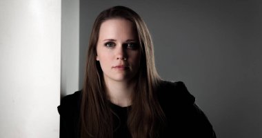 Uzun kahverengi saçlı, siyah gömlekli, beyaz bir duvarın yanında poz veren genç bir kadının stüdyo portresi.