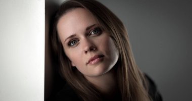 Uzun kahverengi saçlı, beyaz bir duvara yaslanmış düşünceli bir kadının stüdyo portresi.