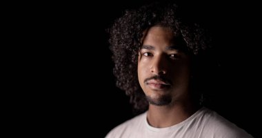 Kıvırcık saçlı, keçi sakallı, beyaz tişörtlü, siyah arka planlı stüdyoda poz veren ciddi bir Afrikalı Amerikalı 'nın yakın portresi.