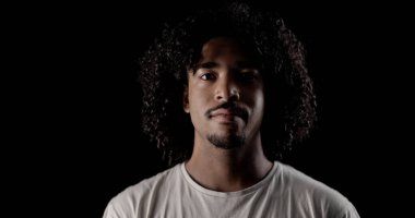 Siyah arka planda kıvırcık saçlı, kendinden emin Afro-Amerikan bir adamın stüdyo portresi.