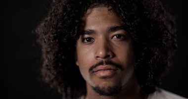 Siyah arka planda poz veren kıvırcık saçlı ve keçi sakallı Afro-Amerikan bir adamın yakın portresi.