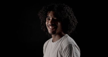 Siyah arka planda günlük tişört giyen kıvırcık saçlı, gülümseyen Afro-Amerikan genç adamın stüdyo portresi.