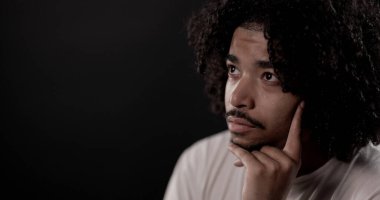 Siyah arka planda kıvırcık saçlı düşünceli genç bir adamın portresi, çenesini tutuyor, başka tarafa bakıyor.