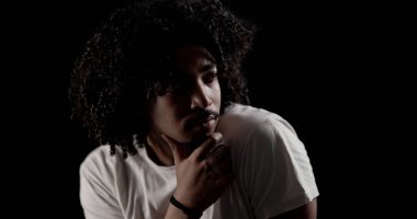 Düşünceli, kıvırcık saçlı ve siyah arka planda çenesine dokunan Afrikalı genç bir Amerikalının stüdyo portresi.