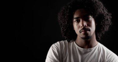 Stüdyoda siyah arka planda poz veren kıvırcık saçlı, ciddi bir Afrikalı Amerikalı adamın portresi.