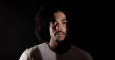 Kıvırcık saçlı ve sakallı, hafif bir kaynakla kısmen aydınlatılmış, ciddi bir Afrikalı Amerikalı adamın portresi.