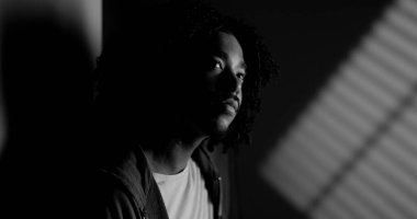 Pencere ışığıyla aydınlatılmış düşünceli bir Afrikalı Amerikalı adamın dramatik siyah beyaz portresi.