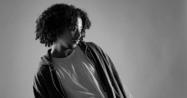 Kıvırcık saçlı, sıradan kıyafetler giyen siyah beyaz bakışlı Afro-Amerikan bir adamın stüdyo portresi.