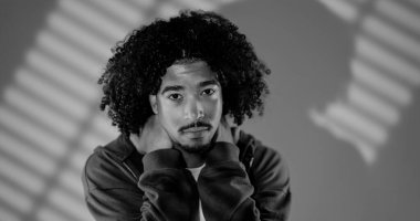 Stüdyoda arka planda çizgili gölgelerle poz verirken boynunu tutan kıvırcık saçlı ciddi bir gencin portresi.