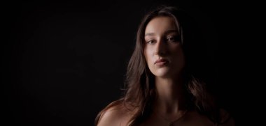 Uzun kahverengi saçlı, koyu bir arka planda çıplak omuzlu, ciddi görünen beyaz bir kadının stüdyo portresi.