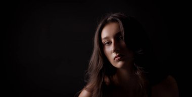 Uzun kahverengi saçlı, siyah arka planda hafif bir ışıkla aydınlatılmış genç bir kadının stüdyo portresi.