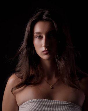 Uzun kahverengi saçlı, beyaz bluz ve kolye giyen genç bir kadının stüdyo portresi.