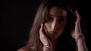 Siyah arka planda yüzüne ve saçlarına bilezikleriyle dokunan çekici genç bir kadının stüdyo portresi.