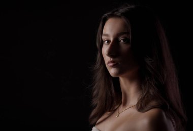 Uzun kahverengi saçlı, narin altın bir kolye takan beyaz bir kadının stüdyo portresi, koyu bir arka fona karşı hafif ışıklandırmayla aydınlatılmış.